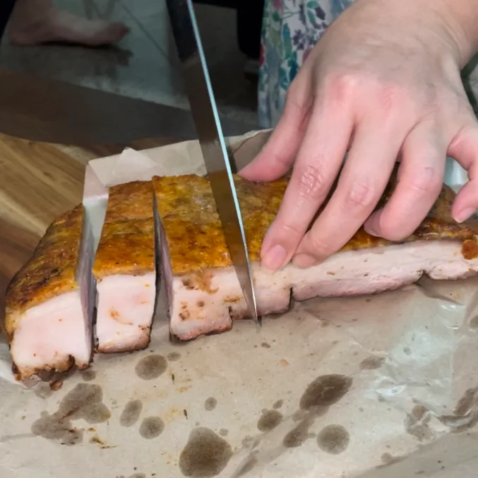 A knife cutting a slab of Happy World Roast roasted pork belly into strips, the crackling splitting under the blade