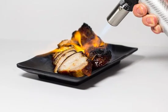 A blowtorch finishing sliced roast pork on a black plate at a Happy World Roast live station