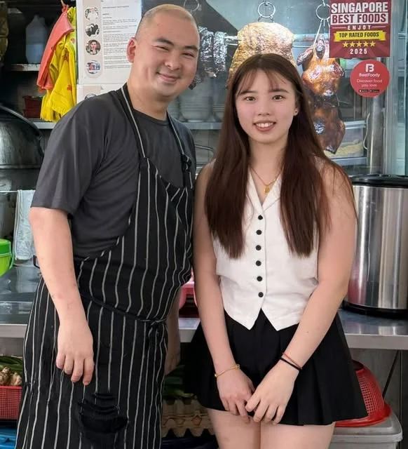 Two people standing at the Happy World Roast counter, roast ducks and chickens hanging on the rail behind them and a Singapore Best Foods Top Rated plaque on the wall